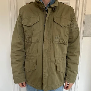 Olivgrön M65 Jacka  - Säljer en olivgrön M65 field jacket med flera praktiska fickor och axelklaffar. Jackan har en dragkedja och knappar framtill samt en hög krage. Perfekt för höst och vår. Storlek M. Se mina övriga annonser för fler grymma plagg! 