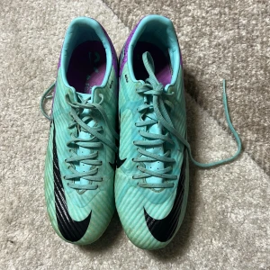 Turkosa fotbollsskor från Nike - Snygga turkosa fotbollsskor från Nike med svarta och lila detaljer. Skorna har snörning och en modern design med en svart Nike-logga på sidan. Perfekta för fotbollsplanen med sina greppvänliga dobbar.