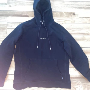 Les Deux hoddie - Säljer en stilren svart hoodie från Les Deux med justerbar huva och känguruficka. Perfekt för en avslappnad look. Den har långa ärmar och en liten logga på bröstet. Passar bra till både jeans och joggers.