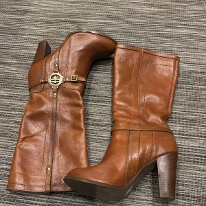 Bruna skinnstövlar med klack - Snygga bruna fordrade stövlar i skinn med hög klack och dekorativa spännen i guld. Perfekta för en stilren look.