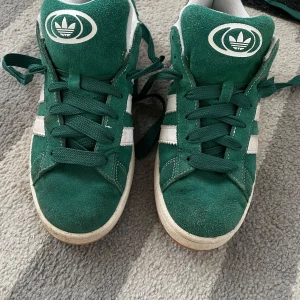 Gröna sneakers från Adidas - Snygga gröna sneakers från Adidas med vita detaljer och klassiska ränder på sidorna. Skorna har snörning och en bekväm passform, perfekt för vardagsbruk.