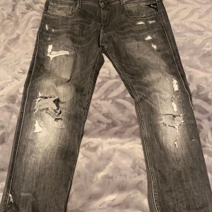 Grå jeans med slitningar - Säljer ett par grå jeans från Replay med coola slitningar och en straight passform. De har klassiska fem fickor och en knappgylf. Perfekta för en avslappnad stil.