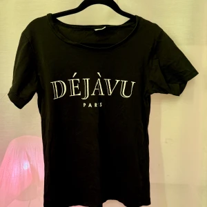 Svart t-shirt med tryck 'Déjàvu Paris' - Snygg svart t-shirt med texten 'Déjàvu Paris' i vitt på framsidan. Perfekt för en casual look. T-shirten har en rund hals och korta ärmar.
