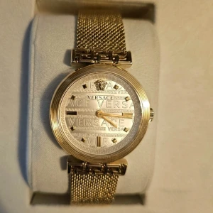 Guldfärgad klocka från Versace - Elegant guldfärgad klocka från Versace med ett detaljerat mönster på urtavlan och ett stilrent mesh-armband. Klockan har en rund boett och tydliga Versace-logotyper på urtavlan. Perfekt för den som vill ha en lyxig accessoar.