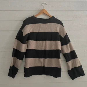 Randig stickad tröja - Säljer en mysig stickad tröja med breda ränder i svart och beige. Tröjan har långa ärmar och en avslappnad passform, perfekt för kyligare dagar. Den är både stilren och bekväm, ett måste i garderoben!