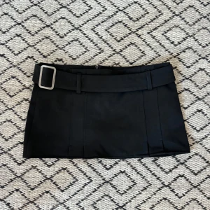 Svart kortkjol med bälte från A feeling of Joy - Snygg svart kortkjol med ett brett bälte och spänne i midjan från A feeling of Joy. Perfekt för att ge en stilren look. Kjolen är enkel och elegant, vilket gör den lätt att matcha med olika toppar. Lowwaist miniskirt. Bältet går även att ta av i midjan! Dragkedja baktill. 