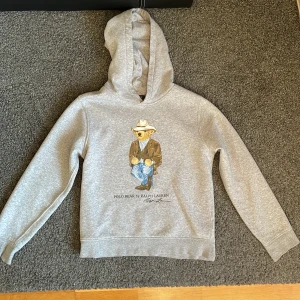 Grå hoodie från Ralph Lauren med Polo Bear - Säljer en grå hoodie från Ralph Lauren med det ikoniska Polo Bear-motivet på framsidan. Tröjan har en klassisk huva.  Tillverkad i mjukt material för extra komfort. Nypris 1699, säljes för 699kr. Inga defekter.