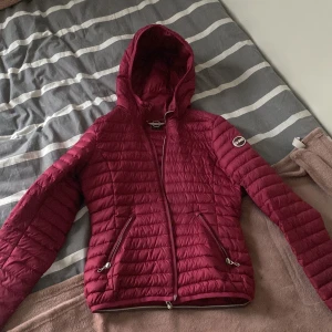 Vinröd dunjacka från Colmar - Snygg vinröd dunjacka från Colmar med huva och dragkedja. Jackan har en quiltad design och två praktiska fickor med dragkedjor. Perfekt för kyligare dagar och ger en stilren look.