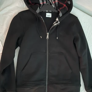 Svart huvtröja från Burberry - Säljer en stilren svart huvtröja från Burberry med dragkedja och rutigt mönster i huvan. Tröjan har långa ärmar och är tillverkad i bomull för en bekväm känsla. Perfekt för en casual look.