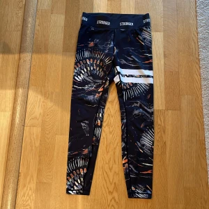 Mönstrade leggings från Stronger - Snygga leggings från Stronger med ett unikt mönster i svart, orange och vitt. De har en hög midja och elastisk linning med Stronger-loggan. Perfekta för träning eller en aktiv vardag.