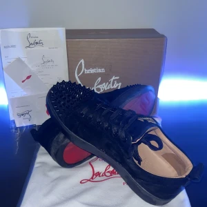 Svarta sneakers från Christian Louboutin - Snygga svarta sneakers från Christian Louboutin med ikoniska röda sulor och nitar på ovansidan. Skorna har snörning och är tillverkade i skinn med en lyxig känsla. Perfekta för att ge en edgy touch till din stil. 1 spik borta. 6,5/10 skick