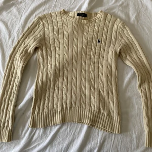 Polo Ralph Lauren Beige - Säljer en stilren beige stickad tröja från Ralph Lauren med kabelstickat mönster och den klassiska loggan broderad i mörkblått på bröstet. Tröjan har långa ärmar och ribbade kanter för en snygg passform. VILL BLI AV MED SNABBT
