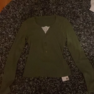 Grön ribbad tröja med spetsdetalj - Säljer en stilren grön ribbad tröja med v-ringning och spetsdetalj vid halsen. Tröjan har långa ärmar och knappar framtill. Perfekt för en chic och bekväm look.