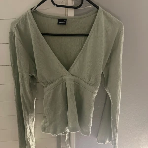 Grön topp från Gina Tricot - Säljer en stilren grön topp från Gina Tricot. Toppen har en v-ringad design och långa ärmar. Den har även ett knytband i midjan för en snygg passform. Perfekt för både vardag och speciella tillfällen. Strl M🩷🩷