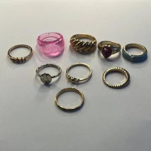 Samling av ringar i olika stilar - En samling av tio ringar i olika stilar och material. Inkluderar en rosa plastig ring, flera guldfärgade ringar och en med en röd sten. Perfekt för att mixa och matcha för olika tillfällen. Går att köpa enskilda också.