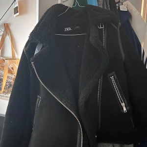 Svart jacka från Zara - Snygg svart jacka från Zara med dragkedja och fuskpälsfoder. Jackan har en cool bikerstil med detaljer som spännen och fickor med dragkedjor. Perfekt för att hålla sig varm och stilren under kyliga dagar.