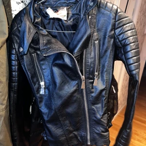 Svart skinnjacka med dragkedjor - Snygg svart skinnjacka med coola dragkedjedetaljer och quiltade axlar. Perfekt för en edgy look. Jackan har en klassisk krage och flera fickor med dragkedjor framtill.