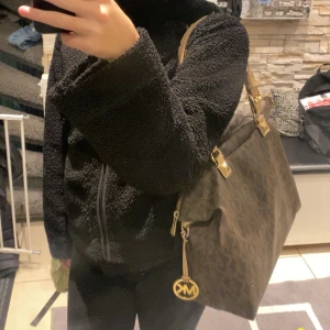 Brun michael kors väska  - Säljer en snygg brun axelväska från michael kors som har guldfärgade detaljer. Väskan har dubbla handtag och en praktisk dragkedja för säker förvaring. Perfekt för att bära med sig det nödvändigaste med stil.