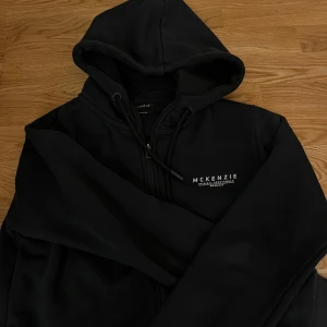 Svart hoodie från McKenzie - Säljer en svart hoodie från McKenzie med dragkedja och huva. Perfekt för en avslappnad stil. Den har en diskret logga på bröstet och är gjord i mjukt material.