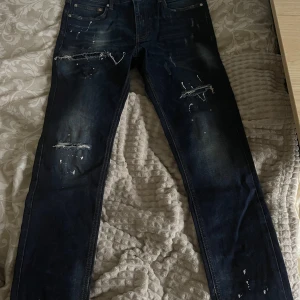 Supply & Demand - Säljer ett par mörkblå jeans med coola slitningar och färgstänk. De har en slim passform och är perfekta för en avslappnad stil. Jeansen är i bra skick och passar både till vardags och fest. Perfekt för dig som vill ha en edgy look!