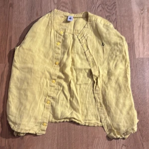 Gul linneskjorta med knappar - Säljer en solgul linneskjorta med knappar framtill. Skjortan har långa ärmar och en avslappnad passform, perfekt för vår och sommar. Den är i bra skick och har en liten broderad detalj på bröstet. 