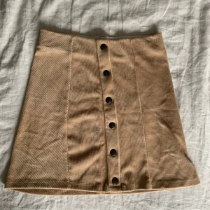 Beige manchesterkjol med knappar - Säljer en superfin beige manchesterkjol i kort modell. Kjolen har snygga knappar framtill som ger en cool look. Perfekt för höst och vår, och kan stylas med både sneakers och boots. Den är i bra skick och passar perfekt för en casual dag ute eller en fika med vänner.