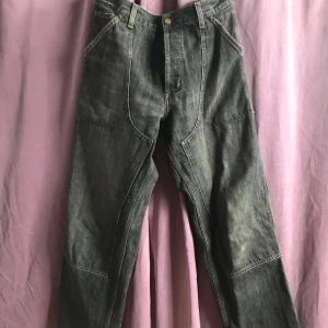 Grå arbetsbyxor från Carhartt - Säljer ett par grå arbetsbyxor från Carhartt i bra skick. Byxorna har en loose passform och är tillverkade i slitstarkt jeansmaterial. De har flera praktiska fickor och en klassisk design med knappar och dragkedja. Perfekta för både arbete och vardag! Mått: 32 x 34