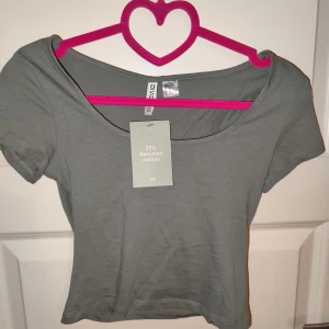 Grågrön kortärmad topp från H&M Divided - Säljer en stilren topp från H&M Divided i storlek M. Den är tillverkad av 25% återvunnen bomull och har en enkel design med korta ärmar och rund hals. Perfekt för vardagsbruk eller att matcha med en snygg jacka.