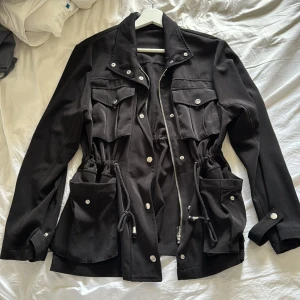 Svart kappa från BikBok - Säljer en svart jacka med dragkedja och knappar. Den har ett snörs  i midjan för en justerbar passform och flera fickor fram. Perfekt för höst och vår! Passar både XS/S då den är lite oversized