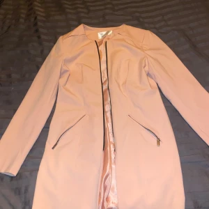 Ljusrosa kappa med dragkedja - Säljer en superfin ljusrosa kappa i mycket bra skick. Den har en stilren design med dragkedja framtill och två snedställda fickor med dragkedjor. Perfekt för vår och höst när du vill vara både snygg och bekväm. Passar till både vardag och fest!