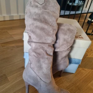 Beige mockastövlar med klack - Snygga beige stövlar i mocka med hög klack och spetsig tå. Perfekta för höst och vinter, dessa stövlar ger en elegant touch till vilken outfit som helst. De har en stilren design utan snörning eller dragkedja, vilket gör dem lätta att ta på och av.