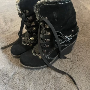 Svarta kilklackade boots från Replay - Snygga svarta boots med kilklack från Replay. De har en mysig pälsfodring och snörning framtill. Perfekta för höst och vinter med sin varma design. Skorna har även en cool broderad detalj på sidan och en robust sula för bra grepp.