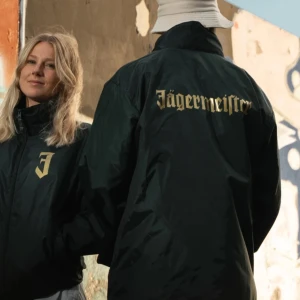 Grön vindjacka från Jägermeister - Säljer en grön vindjacka från Jägermeister med guldtryck. Jackan har en dragkedja framtill och en huva för extra skydd mot väder. Perfekt för höst och vår när det blåser lite extra. Den är i mycket bra skick och har en cool stil med Jägermeister-loggan på ryggen och bröstet.