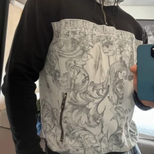 Svart och grå Versace hoodie med mönster - Säljer en snygg svart och grå Versace hoodie med ett detaljerat mönster på framsidan och baksidan. Den har en dragkedja på sidan och en stor huva. Perfekt för höst och vår, och den är i mycket bra skick. Passar både till vardags och lite mer uppklädda tillfällen.