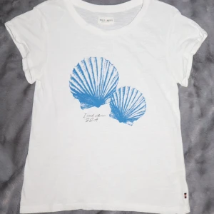 Vit t-shirt med blå snäckmotiv - Vit t-shirt med ett blått snäckmotiv och texten 'I need vitamin SEA'. T-shirten är kortärmad, gjord i bomull och har uppvikta ärmar. Den är i bra skick!