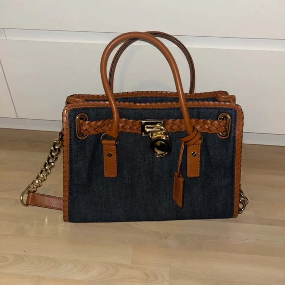 Snygg jeansväska från Michael Kors med flätade bruna läderdetaljer och guldfärgad låsdetalj. Väskan har en kedja som axelrem och ett rymligt innerfack med mönstrat foder. Perfekt för både vardag och fest!. Väskor.