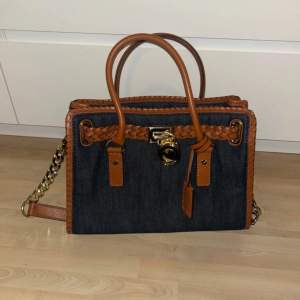 Snygg jeansväska från Michael Kors med flätade bruna läderdetaljer och guldfärgad låsdetalj. Väskan har en kedja som axelrem och ett rymligt innerfack med mönstrat foder. Perfekt för både vardag och fest!