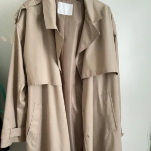 Beige trenchcoat från Samsoe & Samsoe - Säljer en stilren beige trenchcoat från Samsoe & Samsoe.  Jackan är väldigt lång och i lite regn Jack material skulle jag säga. Super fin, älskar den men den kommer tyvärr inte till användning. Köpt på Ullared för 500/699kr. Helt oanvänd 