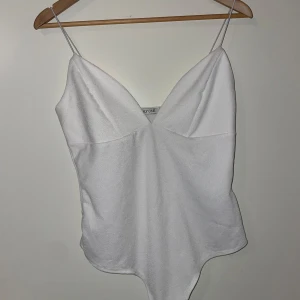Vit ärmlös bodysuit - Säljer en stilren vit bodysuit med smala axelband och v-ringning. Perfekt för sommardagar eller som basplagg under en kavaj. Materialet är mjukt och stretchigt för en bekväm passform. Passar både till vardags och fest!