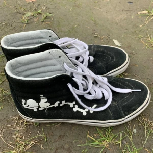 Svarta Vans sneakers med Peanuts - Säljer ett par coola svarta Vans sneakers med vitt tryck av peanuts och  på sidan. Skorna har vita snören och den klassiska 'Off the Wall'-loggan på hälen. Perfekta för en avslappnad stil och passar till det mesta. Använda men i bra skick!