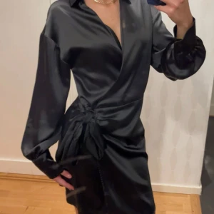 Svart omlottklänning i satin - Snygg svart omlottklänning i satin med långa ärmar och knytning i sidan. Perfekt för en festlig kväll eller en elegant middag. Klänningen har en v-ringad design och ger en lyxig känsla. Passformen är normal och materialet känns mjukt och följsamt mot huden.