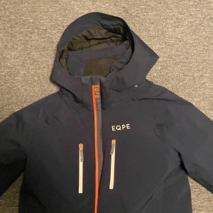Mörkblå jacka från EQPE - Säljer en mörkblå skidjacka från EQPE i mycket bra skick. Den har en praktisk dragkedja i orange och är utrustad med RECCO-teknologi för extra säkerhet. Perfekt för vintern med sin skyddande huva och stilrena design. Passar ungdomar i storlek 158-164.