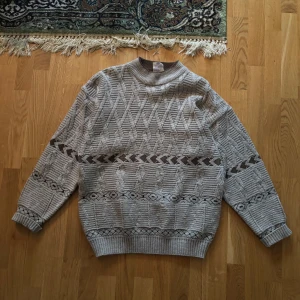 Grå stickad tröja från New Tait - Säljer en grå stickad tröja från New Tait med ett unikt geometriskt mönster. Tröjan har långa ärmar och ribbade muddar vid ärmslut och nederkant. Perfekt för kyliga dagar!