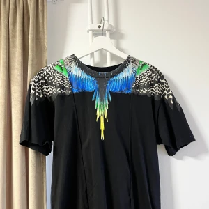 Svart t-shirt med färgglatt mönster från Marcelo Burlon - Snygg svart t-shirt från Marcelo Burlon med ett iögonfallande mönster av färgglada fjädrar i blått, grönt och gult runt halsen. Perfekt för att ge din outfit en unik touch.
