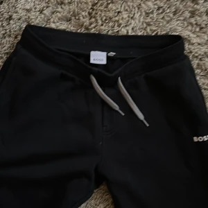 Svarta mjukisbyxor från Boss - Säljer ett par svarta mjukisbyxor från Boss med snörning i midjan. Perfekta för en avslappnad stil. Byxorna har en enkel design med elastiska muddar vid bensluten. Nypris 899:-