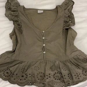 Olivgrön blus från Jacqueline de Yong - Säljer en söt olivgrön blus från Jacqueline de Yong med volangdetaljer och knappar framtill. Den är köpt second hand men jag har aldrig använt den. Blusen har en v-ringad design och är ärmlös med broderade detaljer längs kanterna. Perfekt för en somrig look!