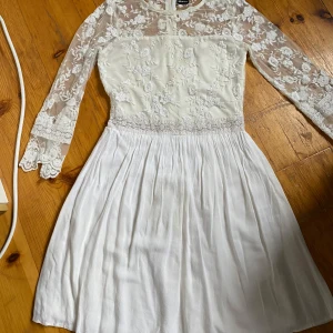 Vit spetsklänning med lång ärm - Elegant vit klänning för studenten  med spetsdetaljer och långärmad design. Klänningen har en vacker blommig spets upptill och en mjukt fallande kjol. Perfekt för speciella tillfällen.