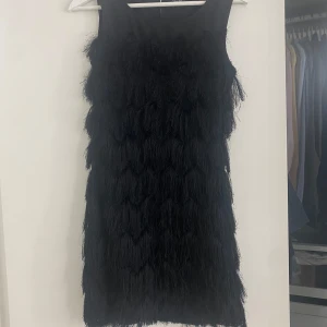 Svart klänning med fransar - Snygg svart ärmlös klänning med fransar som ger en cool och trendig look. Perfekt för en utekväll eller fest. Klänningen har en rak passform och är knälång.