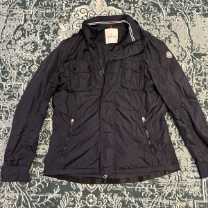 Moncler windbreaker - Säljer en snygg marinblå vindjacka från Moncler i mycket bra skick. Jackan har en stilren design med dragkedja och knappar framtill samt praktiska fickor med lock. Perfekt för höst och vår. Passar både till vardags och lite finare tillfällen.