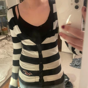 Randig kofta från Hollister - Snygg svartvit randig kofta från Hollister med knappar framtill. Den har långa ärmar och en liten logga på ena sidan.